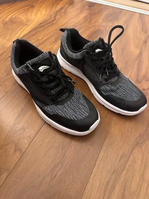 Svarta och grå sneakers med vit sula - Snygga sneakers i svart och grått med vit sula. Ovandelen är i mesh-tyg med svarta detaljer och svarta skosnören. Skorna har en sportig look och är låga i modellen. Perfekta för dig som gillar en clean och stilren stil.
