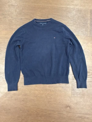 Mörkblå stickad tröja Tommy Hilfiger - Snygg mörkblå stickad tröja från Tommy Hilfiger med rund halsringning och diskret logga på bröstet. Tröjan har ribbade muddar vid ärmslut och nederkant samt en klassisk passform. Perfekt till jeans eller kjol för en clean look.