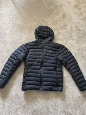 Svart dunjacka med huva - Snygg svart dunjacka med huva och dragkedja framtill. Jackan har två sidofickor med dragkedja och quiltad design med horisontella sömmar. Perfekt för kalla vinterdagar och ger en stilren look.