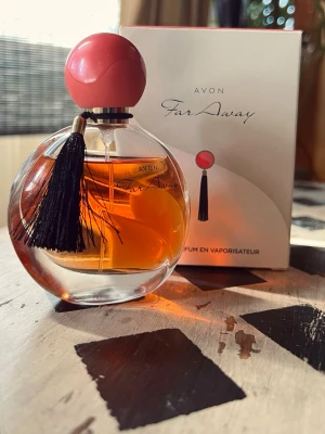 Avon Far Away parfym, 50ml - Avon Far Away parfym i rund glasflaska med orange doftvätska. Flaskan har en stor rosa kork och dekoreras med en svart tofs. Förpackningen är elegant med matchande design och text. Perfekt för dig som gillar klassiska och feminina dofter. Ny, bara testad. 
