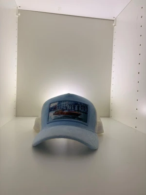 Ljusblå meshkeps med tryck - Säljer en ljusblå keps med vit mesh baktill och böjd skärm. Framtill finns ett tryck med motiv av en båt och byggnader. Kepsen har justerbar snapback och är tillverkad i bomull och polyester. Perfekt för en avslappnad streetwear-look.