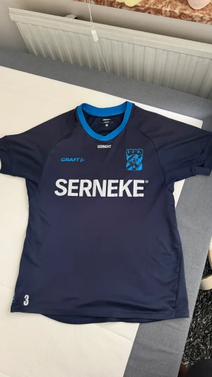 Blå IFK Göteborg matchtröja Craft - Snygg mörkblå matchtröja från IFK Göteborgs A-lag med ljusblå detaljer och klubbmärke på bröstet. Tröjan har rund halsringning och stora vita tryck med sponsorlogga på framsidan. Perfekt för fotbollsträning eller att bära på läktaren.
