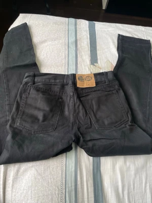 Svarta raka jeans med läderpatch - Säljer ett par svarta jeans med rak passform och klassisk femficksdesign. Jeansen har en snygg läderpatch bak i midjan och är tillverkade i ett slitstarkt bomullsmaterial. Perfekta för dig som gillar en enkel och stilren look.