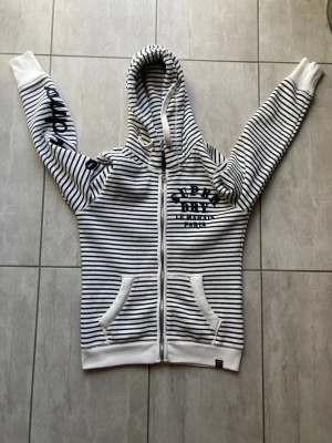 Superdry hoodie - Snygg vit hoodie med svarta horisontella ränder och dragkedja framtill. Tryck på bröstet och ena ärmen, samt två fickor fram. Huva med snörning och ribbade muddar. Perfekt för en chill och avslappnad stil. Vid frågor och funderingar är det bara att höra av sig. Storlek S (står M i nacken men är som S)