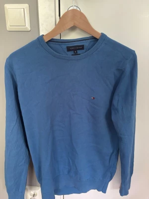 Blå tröja från Tommy Hilfiger - Snygg blå rundhalsad tröja från Tommy Hilfiger i premium bomull. Tröjan har en liten broderad logga på bröstet och ribbade muddar vid ärmslut och nederkant. Perfekt för dig som gillar klassisk och stilren design. Tröjan är i ett mycket bra skick och har inga defekter eller liknande, hör av er vid frågor!