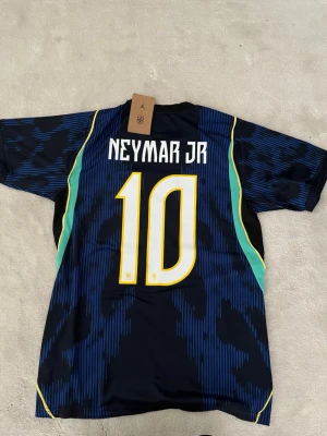 Brasilien Neymar Jr tröja - Snygg blå matchtröja från Brasilien med Neymar Jr och nummer 10 på ryggen. Tröjan har gula och gröna detaljer, rund hals och korta ärmar. Tillverkad i ett lätt och ventilerande material, perfekt för fotboll eller träning.