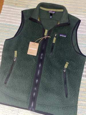 Patagonia grön väst XS - Mörkgrön fleeceväst från Patagonia med dragkedja framtill. Västen har tre fickor med dragkedja och detaljer i olivgrönt tyg. Perfekt lager för kyliga dagar och outdoor-stil. Inga dekekter alls, använd några enstaka gånger! Skriv om du undrar något!