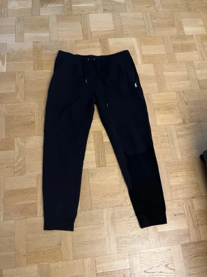 Svarta mjukisbyxor från Ralph lauren  - Säljer ett par svarta mjukisbyxor med dragsko i midjan och ribbade muddar vid bensluten. Byxorna har en liten vit logga på vänster sida framtill och är tillverkade i ett mjukt och bekvämt material, perfekt för chill dagar.