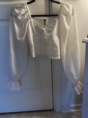 Vit långärmad blus från FB Sister - Söt vit blus från FB Sister med långa puffärmar och resår vid ärmslut. Blusen har en smockad struktur, knytning framtill och en lätt croppad passform. Perfekt för dig som gillar romantisk och trendig stil.