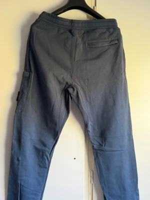 Blå mjukisbyxor från Stone Island - Säljer ett par blå mjukisbyxor från Stone Island i storlek M. Byxorna har en bakficka med dragkedja och en sidoficka med klassisk Stone Island-patch. Sköna och stilrena, perfekta för chill eller streetwear.