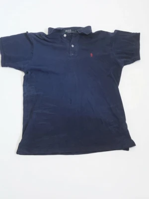 Marinblå pikétröja från Polo Ralph Lauren - Klassisk marinblå pikétröja från Polo Ralph Lauren med korta ärmar och krage. Tröjan har två knappar vid halsen och den ikoniska röda broderade loggan på bröstet. Tillverkad i mjuk bomull för en bekväm känsla.