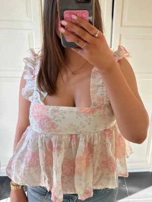Blommig volangblus med öppen rygg - Supersöt blus med volangdetaljer och blommigt mönster i ljusa rosa och vita toner. Toppen har fyrkantig ringning, knyt i ryggen och är ärmlös med breda axelband. Perfekt för sommarens alla tillfällen. Använd ca 1-2 gånger💕