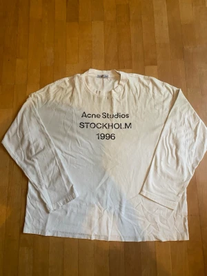 Vit långärmad Acne Studios t-shirt - Vit långärmad t-shirt från Acne Studios med tryck på bröstet där det står 'Acne Studios STOCKHOLM 1996'. Klassisk rund hals och relaxed fit. Perfekt för dig som gillar minimalistisk och stilren design.