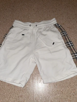 Vita shorts från Berberg med rutiga sidopaneler - Snygga vita shorts med klassiska rutiga sidopaneler i beige, svart och rött. Shortsen har resårmidja med snörning och en bakficka. Perfekta för sommaren och ger en trendig look.