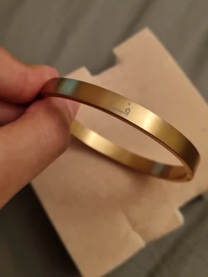 Guldfärgat stelt armband med gravyr - Snyggt stelt armband i guldfärgad metall med en enkel gravyr på utsidan. Armbandet har en minimalistisk och stilren design som passar perfekt till din vardagsstil. Lätt att matcha med andra smycken och accessoarer.