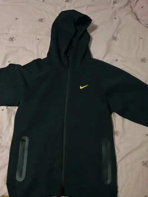 Nike nochta fleece - Storlek L. Säljer en svart hoodie från Nike med dragkedja och huva. Framtill finns en gul Nike-logga och två fickor med dragkedja. På baksidan finns små gula detaljer och text. Hoodien har en sportig look och är tillverkad i ett mjukt material.
