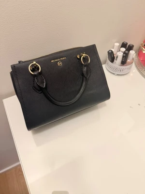Svart handväska från Michael Kors - Snygg svart handväska från Michael Kors i skinn med guldfärgade metalldetaljer och dubbla handtag. Väskan har flera fack och dragkedja upptill, perfekt för att organisera dina grejer. Klassisk och stilren design som passar till det mesta.