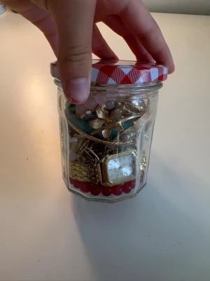 Mystery jewellery jar - En glasburk fylld med olika smycken som armband, halsband och klocka. Smyckena är i flera färger som guld, turkos, rosa och röd. Innehåller pärlor, metallkedjor och dekorativa detaljer. Perfekt för dig som gillar att mixa och matcha accessoarer.