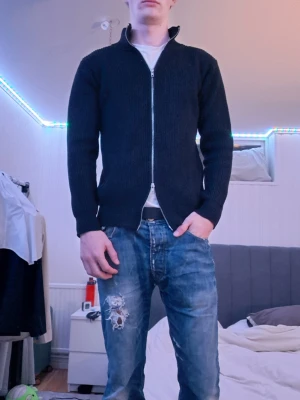 Svart ribbad ziptröja med hög krage - Säljer en omsydd svart ribbstickad ziptröja med hög krage och hel dragkedja framtill. Tröjan har en clean look och är tillverkad i ett tjockare material som passar perfekt när det är lite kyligare. Enkel att matcha med olika outfits.