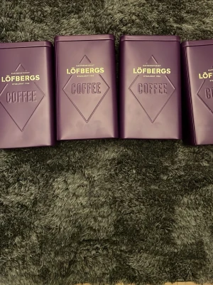 Lila kaffeburk Löfbergs - Säljer fyra  stilrena lila kaffeburkar från Löfbergs med präglad text 'COFFEE' och guldfärgad logga. Burken är rektangulär och tillverkad i metall, perfekt för att förvara kaffe eller andra torrvaror i köket.