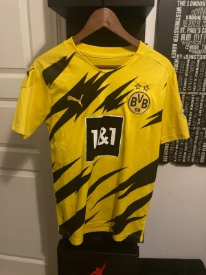 Gul Borussia Dortmund matchtröja Puma - Säljer en gul Borussia Dortmund matchtröja från Puma med svarta blixtmönster och klubbmärke på bröstet. Tröjan har korta ärmar, rund hals och 1&1 sponsortryck på framsidan. Perfekt för dig som älskar fotboll och vill sticka ut på planen.