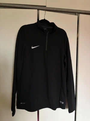 Svart Nike Dri-Fit träningströja - Säljer en svart långärmad träningströja från Nike med Dri-Fit material som transporterar bort fukt. Tröjan har halv dragkedja framtill och en vit Nike-logga på bröstet. 