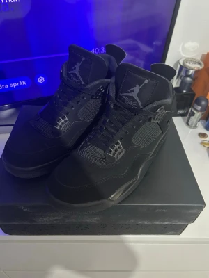 Svarta Air Jordan 4 sneakers - Säljer ett par helsvarta Air Jordan 4 sneakers med klassisk mesh och läderdetaljer. Skorna har högt skaft, ikonisk Jumpman-logga på plösen och häl, samt svarta skosnören. Perfekta för dig som gillar streetwear och vill ha en stilren look.