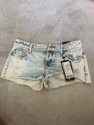 Ljusblå denimshorts med slitningar - Säljer ett par ljusblå denimshorts med slitna detaljer och upprullad kant. Shortsen har klassisk femficksdesign, bälteshällor och knappgylf. Perfekta för varma dagar och ger en avslappnad look.