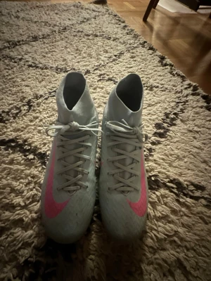 Ljusblå fotbollsskor med rosa swoosh - Snygga fotbollsskor i ljusblått med högt skaft och rosa Nike-swoosh på ovansidan. Skorna har snörning framtill och är designade för bra grepp på planen. Perfekta för dig som vill sticka ut på matchen.