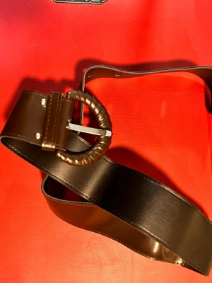 Brett brunt bälte med rund spänne - Snyggt brett bälte i mörkbrunt skinn med ett unikt runt spänne som är lindat i matchande material. Perfekt för att lyfta vilken outfit som helst och ger en cool vintagekänsla. Passar till jeans, kjol eller klänning.