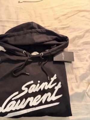 Svart Saint Laurent hoodie med tryck - Svart hoodie från Saint Laurent med vit logotyp i graffiti-stil på bröstet. Klassisk modell med huva och dragsko samt känguruficka framtill. Perfekt för en avslappnad och trendig look.