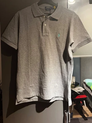 Grå pikétröja från Polo Ralph Lauren - Klassisk grå pikétröja från Polo Ralph Lauren. Tröjan har två knappar vid halsen och den ikoniska broderade loggan i ljusblått på bröstet. Tillverkad i mjuk bomull för en bekväm känsla.