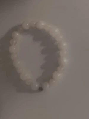 Vitt pärlarmband med rundade pärlor - Snyggt armband med vita, rundade pärlor trädda på elastisk tråd. En av pärlorna är i silverfärg vilket ger en liten kontrast till det annars helvita armbandet. Perfekt accessoar för att lyfta din outfit.