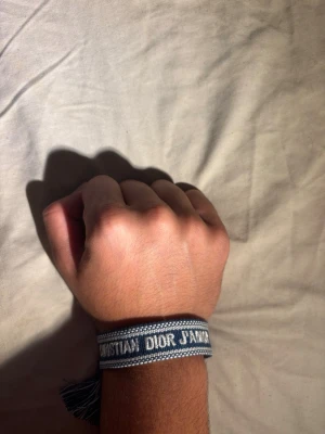 Dior armband - Snyggt blått tygarmband från Christian Dior med texten 'J'ADIOR' invävd i vitt. Armbandet har en bohemisk känsla med tofsar i ändarna och är justerbart för att passa de flesta handleder. Perfekt accessoar för att lyfta din outfit.