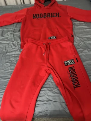 Rött Hoodrich set med huva - Snyggt rött set från Hoodrich med matchande hoodie och byxor. Hoodien har stor huva, magficka och svart logga framtill. Byxorna har dragsko i midjan, fickor och tryckta detaljer med Hoodrich-logga. Perfekt för en avslappnad och trendig look. Priset kan diskuteras vid snabb affär.