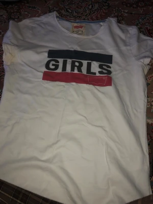 Vit t-shirt med tryck från Levi's - Snygg vit t-shirt från Levi's med trycket 'GIRLS' i svart, blått och rött på bröstet. Klassisk rund halsringning och korta ärmar. Perfekt för en avslappnad och trendig stil.