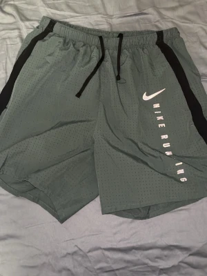 Grå Nike Dri-Fit löparshorts eller badbyxor  - Säljer ett par grå Nike Dri-Fit löparshorts  eller badbyxor, med svarta detaljer längs sidorna och justerbar dragsko i midjan. Shortsen har ventilerande meshmaterial och texten 'NIKE RUNNING' tryckt på ena benet. Perfekta för träning eller löpning.