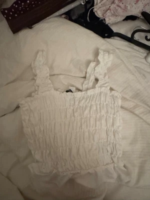 Vit smockad linnetopp med volang - Supersöt vit linnetopp med smockad resår och breda axelband. Toppen har volangkant nedtill och är perfekt för sommarens alla outfits. Tillverkad i ett lätt och luftigt material som känns skönt mot huden.
