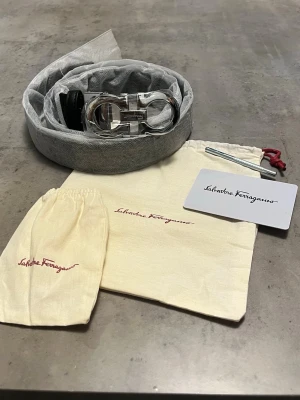 Salvatore Ferragam0 svart skärp - Stilrent svart skärp från Salvatore Ferragam0 med präglat mönster i lädret och en ikonisk silverfärgad spänne. Skärpet levereras med originalpåse, perfekt för att lyfta din outfit med en lyxig touch.                                                      Bältestång medföljer även!