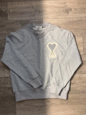 Ami sweatshirt - Säljer en stilren grå sweatshirt med rund hals och ribbade muddar. På bröstet finns en stor applikation med ett hjärta och bokstaven A i vitt och grått. Perfekt för dig som gillar enkel men snygg design.