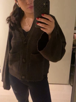 Brun kofta - Mysig brun kofta med v-ringning och stora knappar framtill. Modellen är oversized med breda ärmar och två rymliga fickor fram. Ribbstickade muddar vid ärmslut och nederkant ger en avslappnad look. Perfekt att slänga över en t-shirt eller linne.