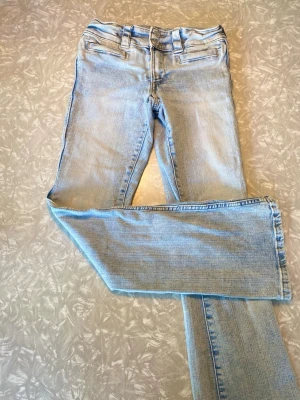 Ljusblå raka jeans med fickdetalj - Säljer ett par ljusblå jeans med raka ben och klassisk femficksdesign. Jeansen har bälteshällor och en snygg fickdetalj framtill. Tillverkade i denim.