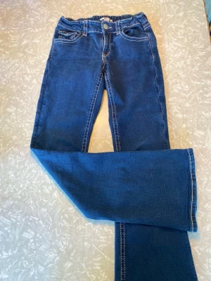 Blå bootcut jeans från Gina Tricot - Säljer ett par blå bootcut jeans från Gina Tricot i storlek 170. Jeansen har klassisk femficksmodell, vita kontrastsömmar och knapp med logga. Materialet är mjukt denim och passformen är utsvängd nedtill. Perfekta för dig som gillar retrostil!