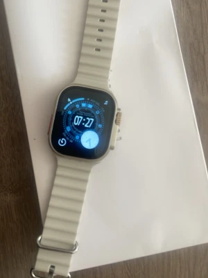 Apple Watch Ultra 3 med vit silikonband 1:1 - Apple Watch Ultra 3 med fyrkantig skärm och vit silikonrem 1:1. Klockan har en robust design med digital krona och orange knapp på sidan. Perfekt för dig som vill ha en smartklocka med många funktioner och modern look.