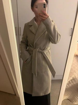 Ljusgrå klassisk kappa med knytskärp - Snygg ljusgrå kappa med bred krage och knytskärp i midjan. Modellen är rak och har diskreta fickor på sidorna. Perfekt för dig som gillar stilrena och tidlösa plagg. Materialet ser ut att vara ullblandning för extra värme.
