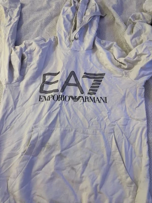 Vit hoodie från Emporio Armani EA7 - Säljer en vit hoodie från Emporio Armani EA7 med stor logga framtill. Hoodien har huva med dragsko och en stor magficka. Tillverkad i mjukt bomullsmaterial som är skönt mot huden. Perfekt för dig som gillar stilrena och exklusiva plagg. Fläckar på magen