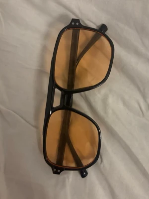 Svarta solglasögon med orange glas - Säljer ett par solglasögon med svart, fyrkantig båge och stora orange tonade glas. Ramen är i plast och har en retroinspirerad design med dubbelbrygga. Perfekt accessoar för dig som vill sticka ut i sommar.