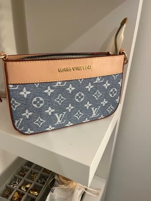 Blå beige denim handväska - Trendig handväska i blått denimtyg med klassiskt monogrammönster och detaljer i ljust läder. Väskan har guldfärgade metalldetaljer, dragkedja upptill och en justerbar axelrem i läder. Perfekt för dig som gillar exklusiv och modern stil.