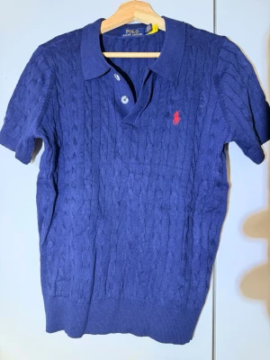 Blå kabelstickad polotopp från Polo Ralph Lauren - Aldrig använt. Toppen har två knappar framtill och den ikoniska röda loggan broderad på bröstet. Perfekt till jeans eller kjol för en stilren look. 