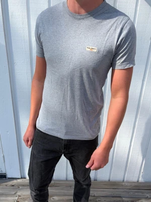 Grå t-shirt från Nudie Jeans - Snygg grå t-shirt från Nudie Jeans med liten logopatch på bröstet. Klassisk rund halsringning och korta ärmar. Tillverkad i mjuk bomull för en skön känsla och enkel stil som funkar till det mesta. Sjukt fet men clean. Bra basic.